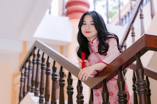 Free Woman in pink Ao Dai holding red fan, leaning on staircase railing indoors. Stock Photo