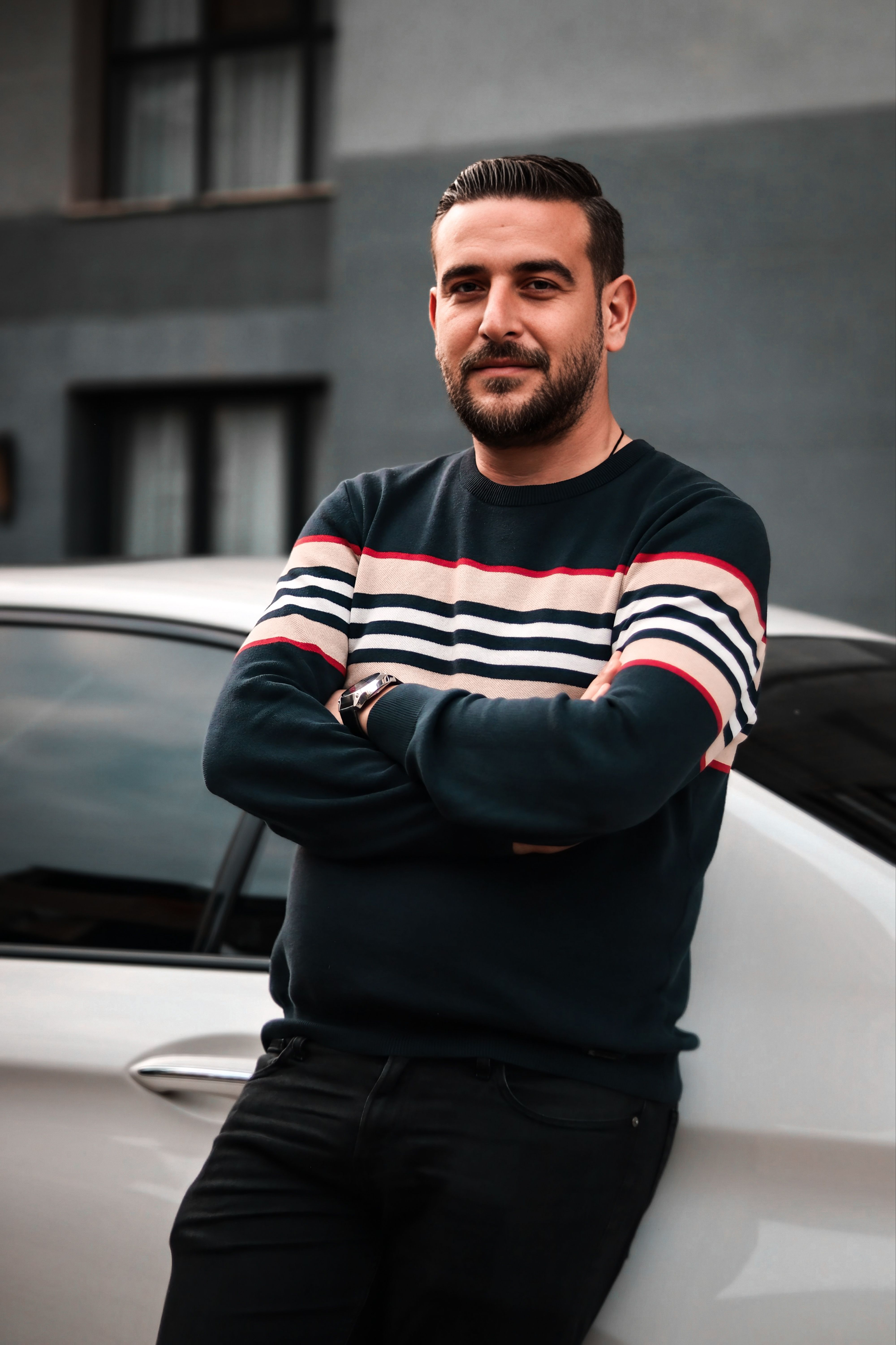 Confident man standing against a car outdoors, showcasing casual style with a striped sweater in a modern urban setting.