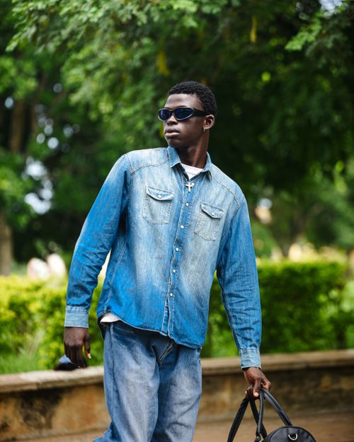 Free Stylish young man wearing denim clothes and sunglasses, carrying a bag in a green park setting. Stock Photo