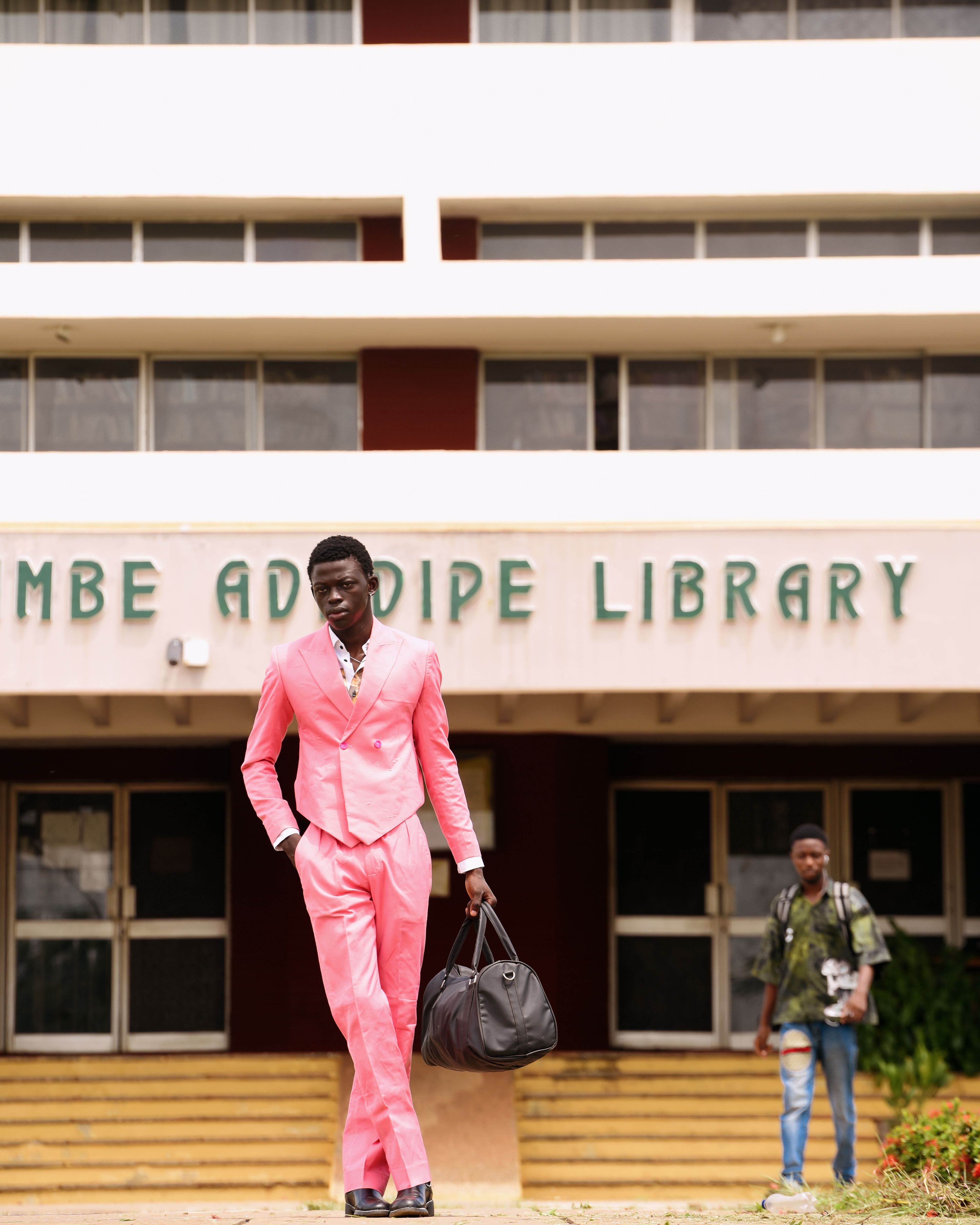A fashionable man in a pink suit stands confidently outside a university library, carrying a black bag.