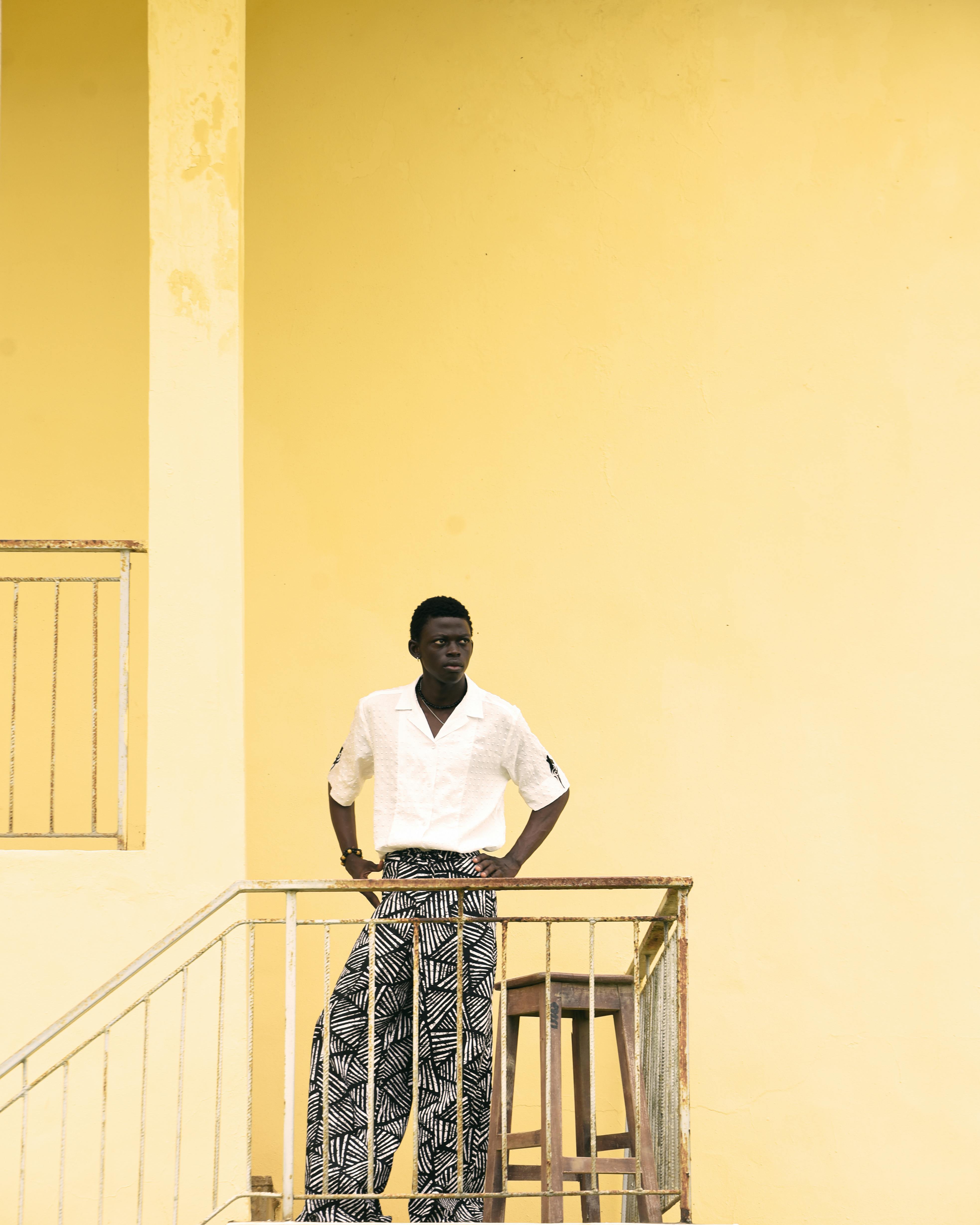 A stylish man in patterned pants pauses by a vibrant yellow wall for a portrait.