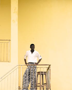 A stylish man in patterned pants pauses by a vibrant yellow wall for a portrait.