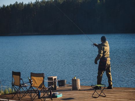 Man fishing on a serene lake pier with chairs, enjoying nature and outdoor tranquility.