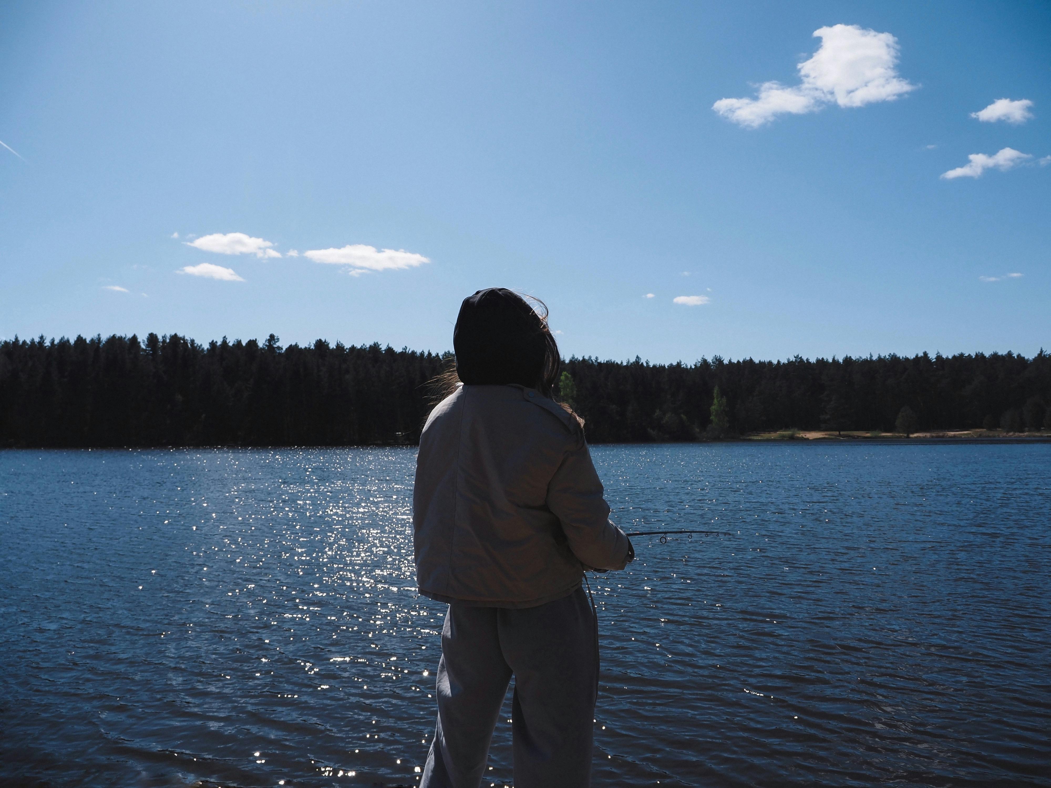 A person enjoys fishing by a calm lake under a bright blue sky, creating a peaceful outdoor scene.
