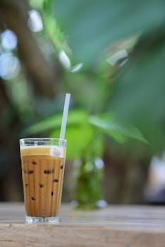 Chilled iced coffee with straw on wooden table against lush green blurred background, perfect for a refreshing summer day.