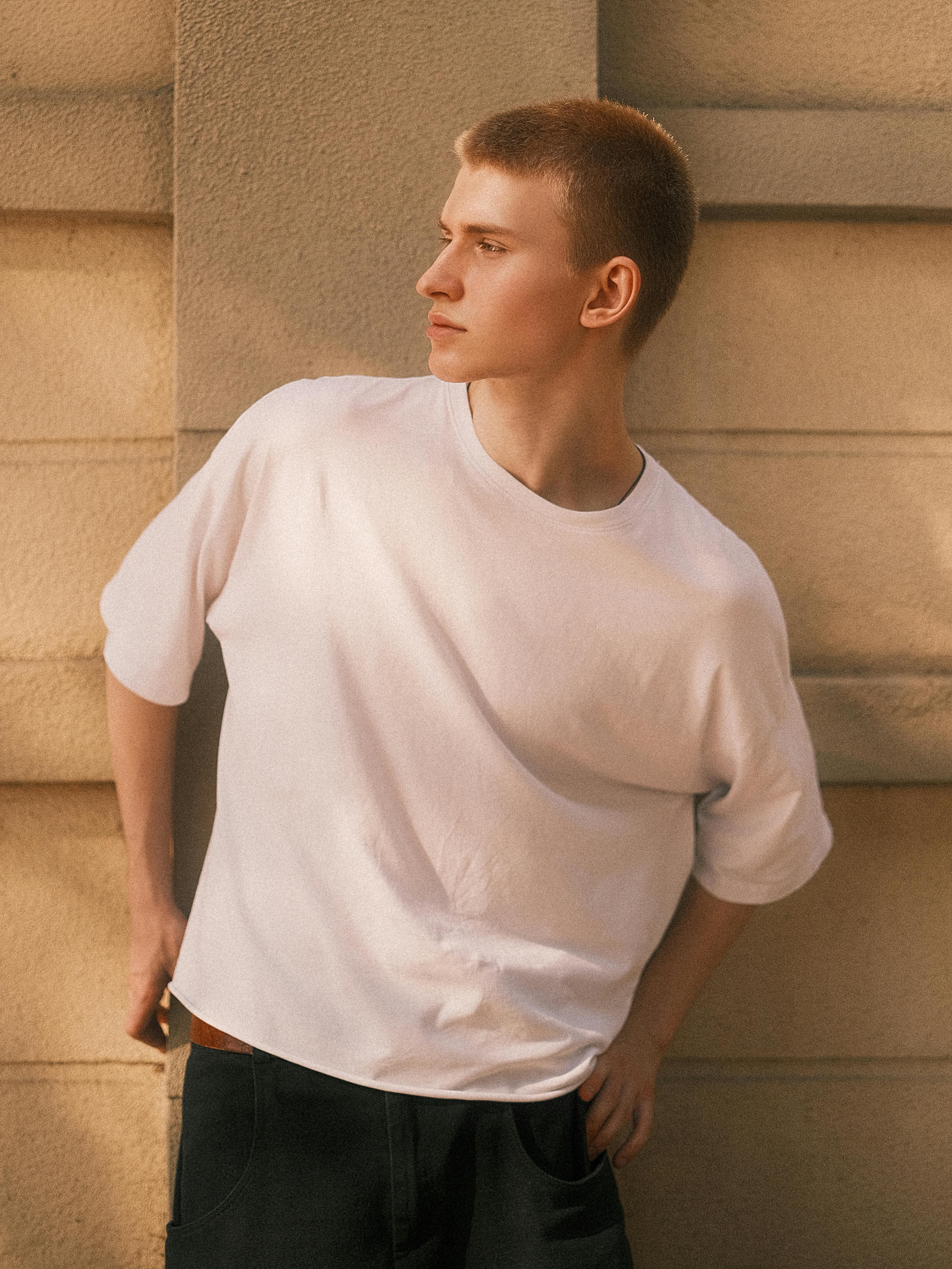 Portrait of a young man in a white t-shirt leaning against a wall with soft lighting.