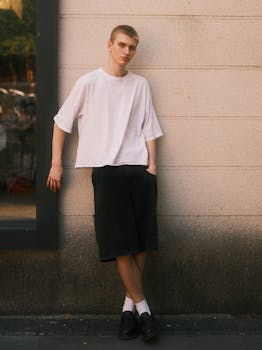 Young adult male in white shirt and black shorts leaning against a wall in urban setting.