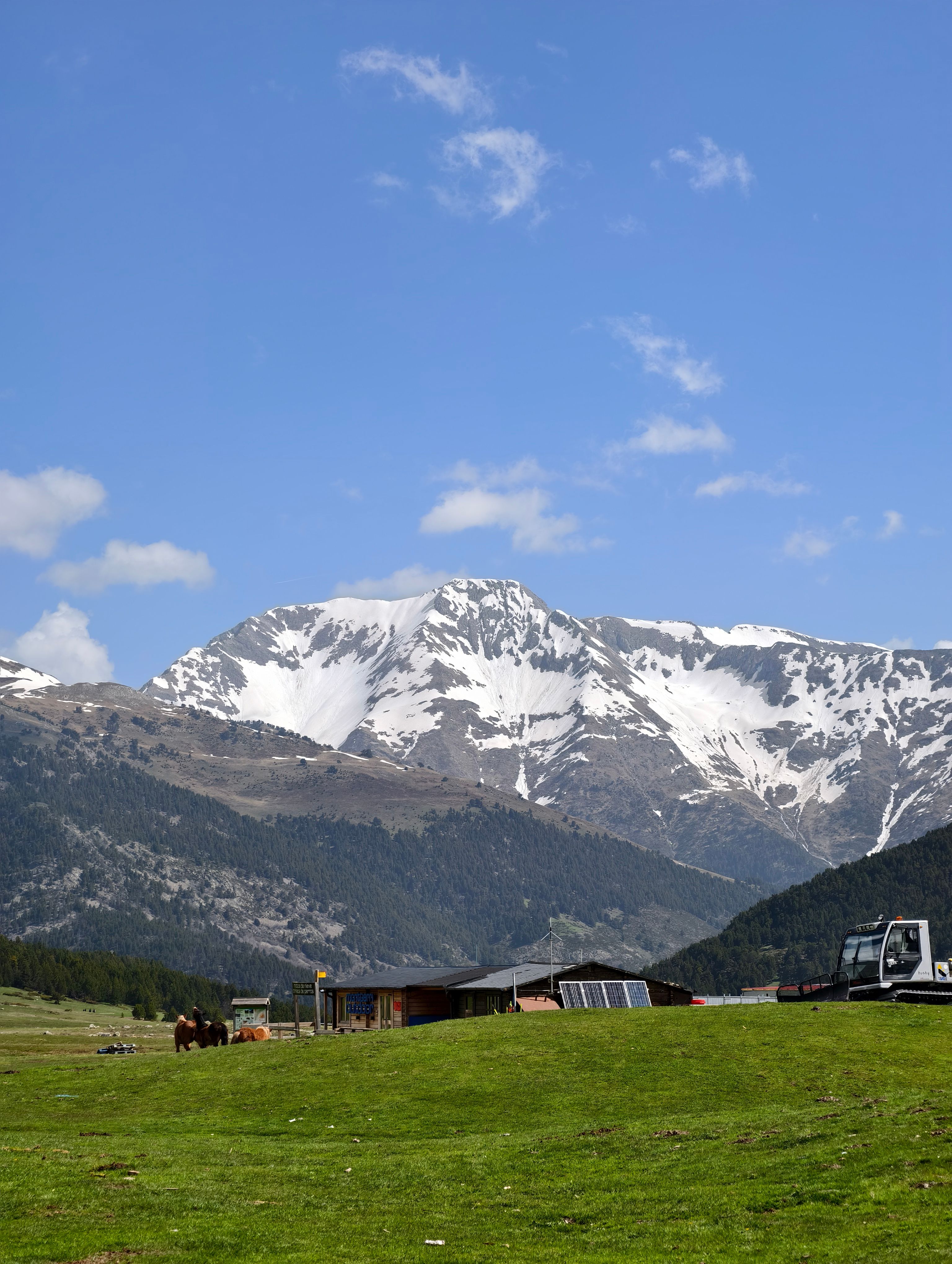 Breathtaking view of a snow-capped mountain range with lush green fields and scattered clouds under a blue sky.