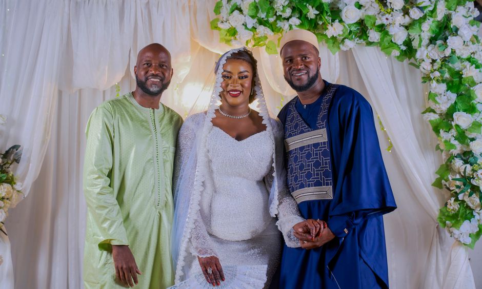 Bride in elegant dress poses with two men in traditional attire amidst floral decorations.