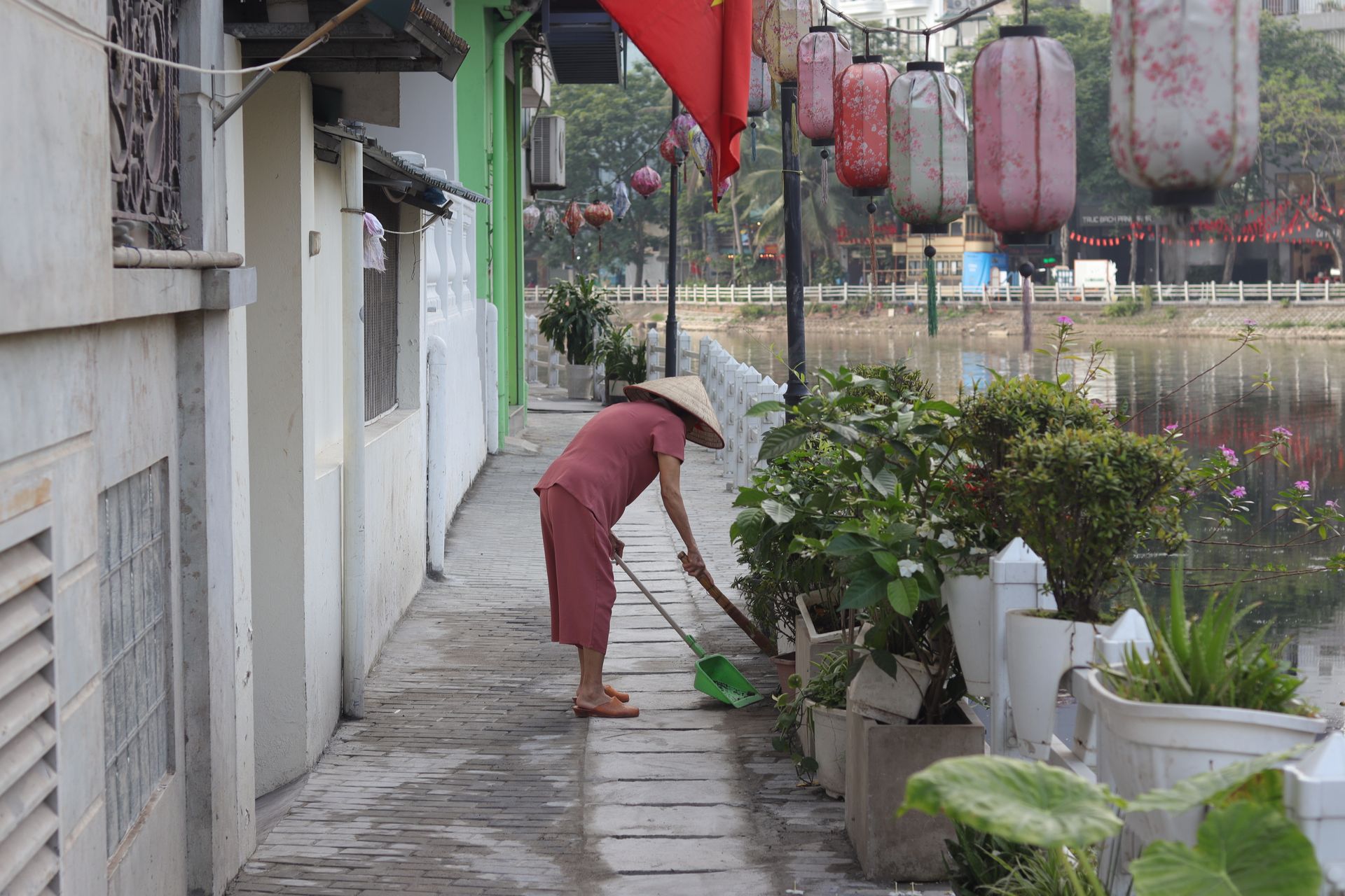 Elderly woman sweeping a riverside path in Hanoi, Vietnam with traditional decorations overhead.