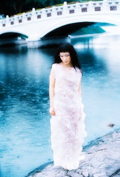 Elegant woman in a white dress stands by a tranquil riverside near a classic white bridge.