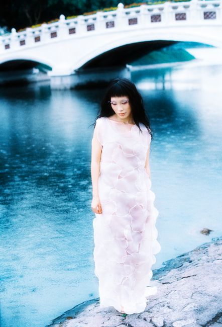 Elegant woman in a white dress stands by a tranquil riverside near a classic white bridge.