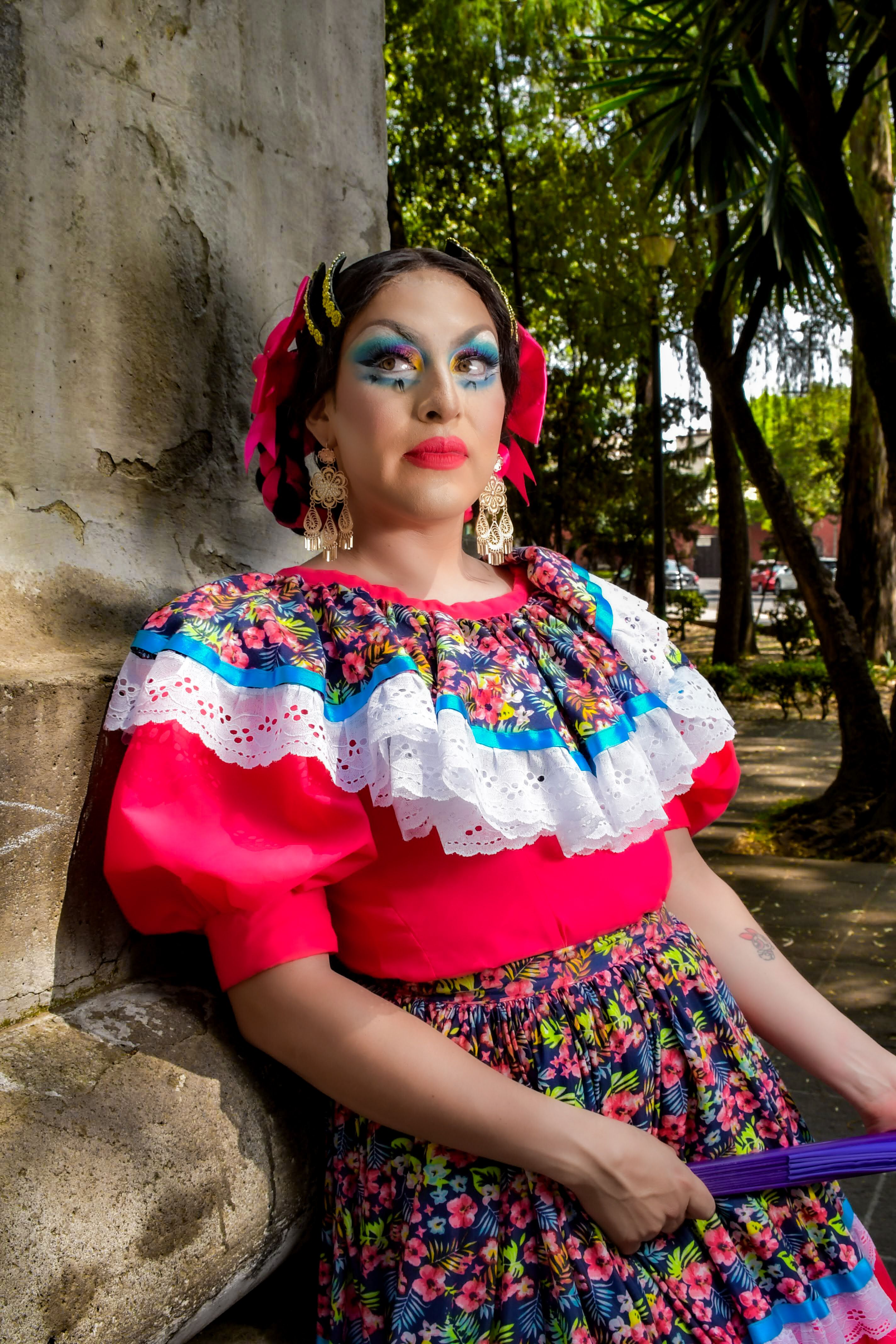 A person in vibrant traditional attire posing outdoors by a wall under sunlight.