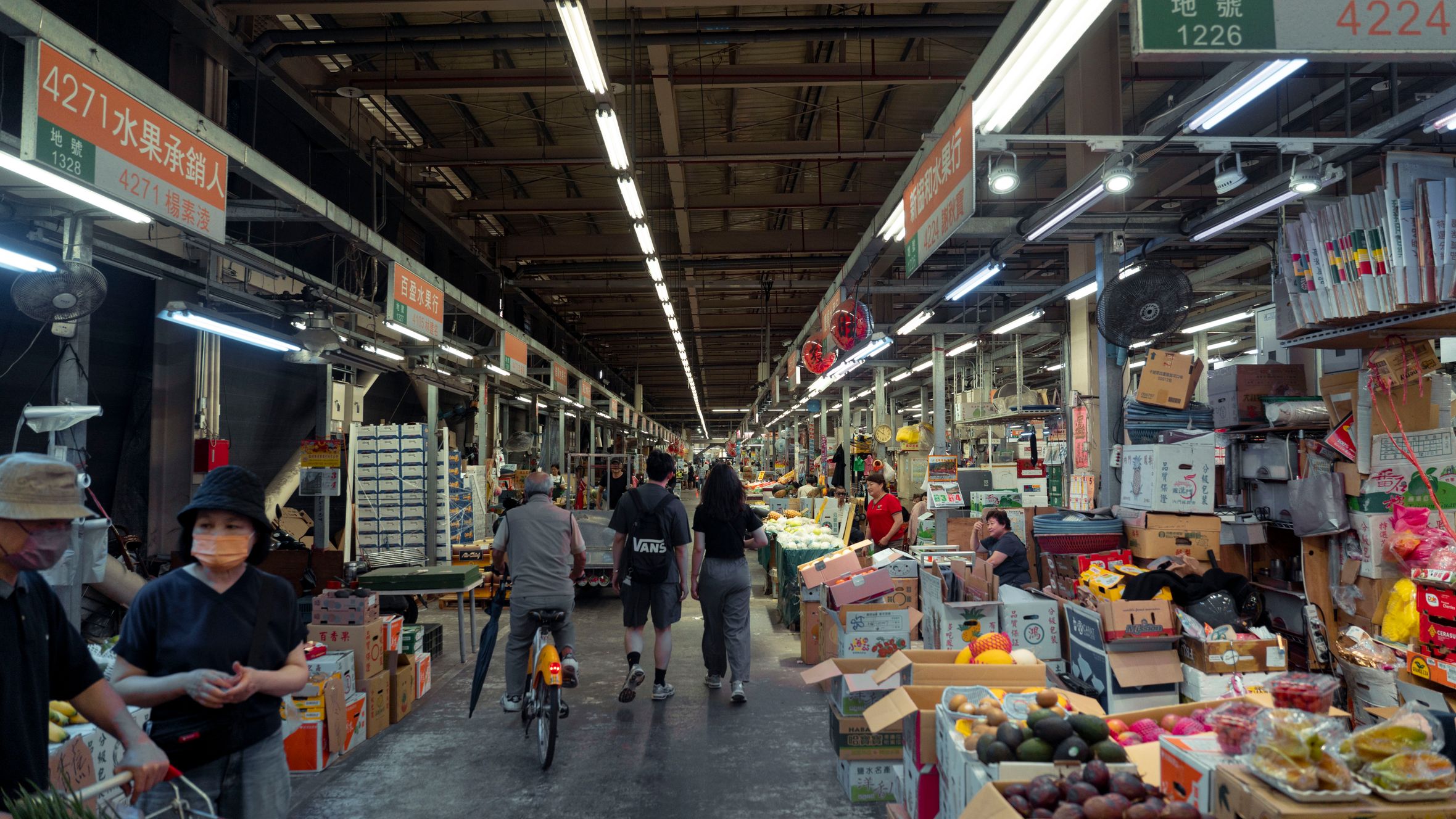 Indoor street market with diverse people and fresh produce displays in an Asian setting.
