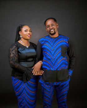Smiling couple in matching blue and black traditional outfits posed on a dark background.