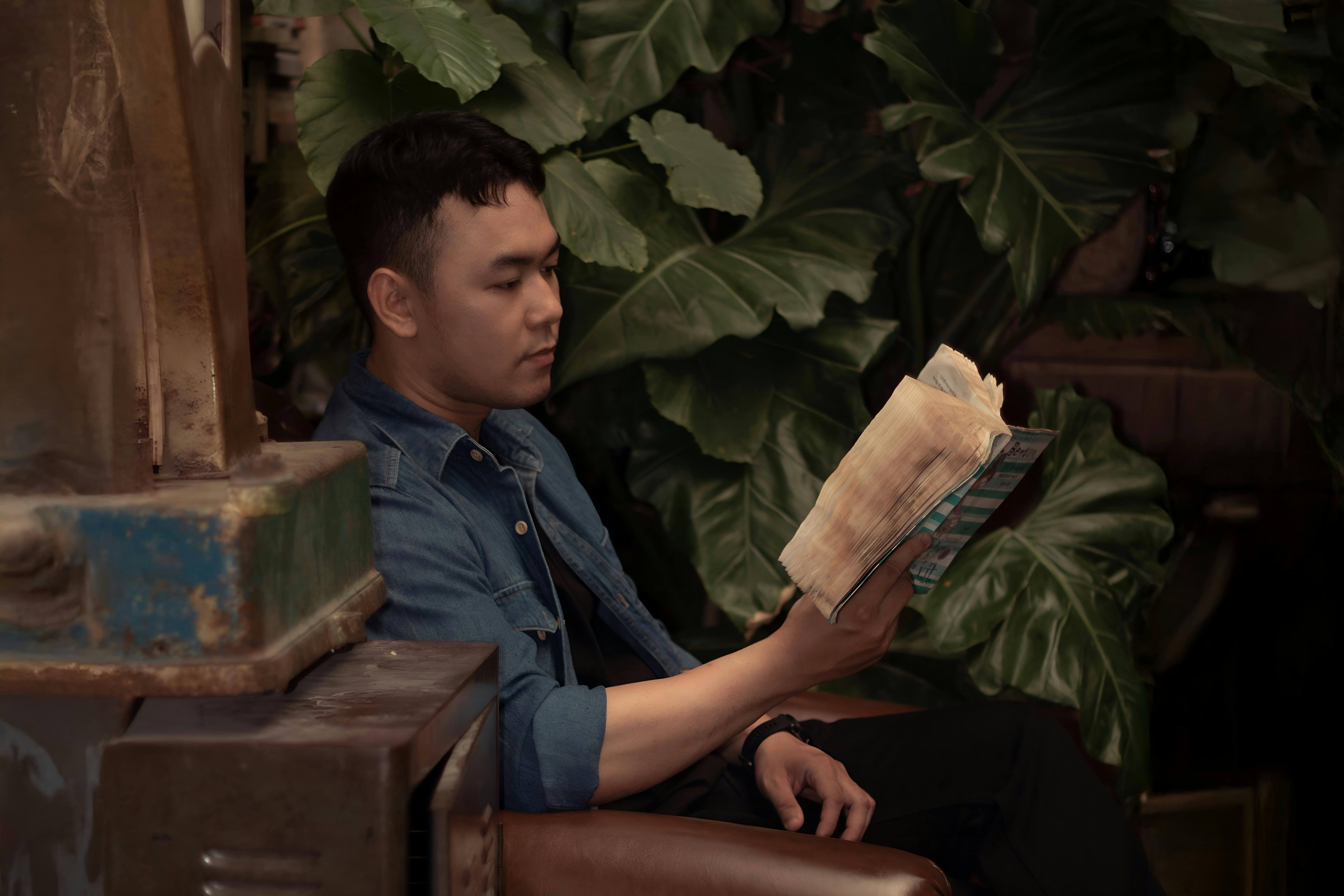 Gratis Seorang pemuda yang tengah asyik membaca buku, dikelilingi tanaman hijau subur di dalam ruangan. Foto Stok