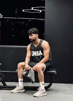 Young man taking a break on gym bench wearing basketball attire, towel in hand.