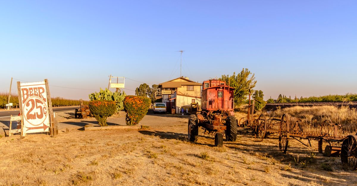 A rustic scene with an old tractor, vintage beer sign, and rural landscape under clear skies.