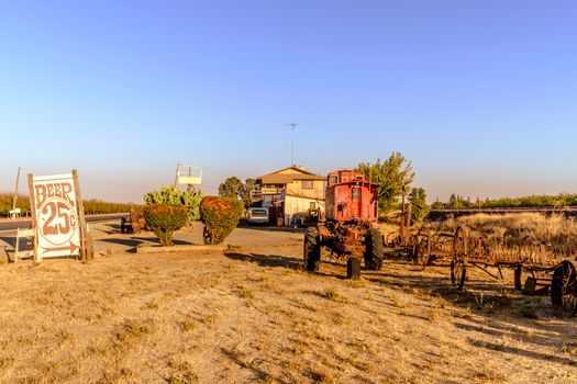 A rustic scene with an old tractor, vintage beer sign, and rural landscape under clear skies.