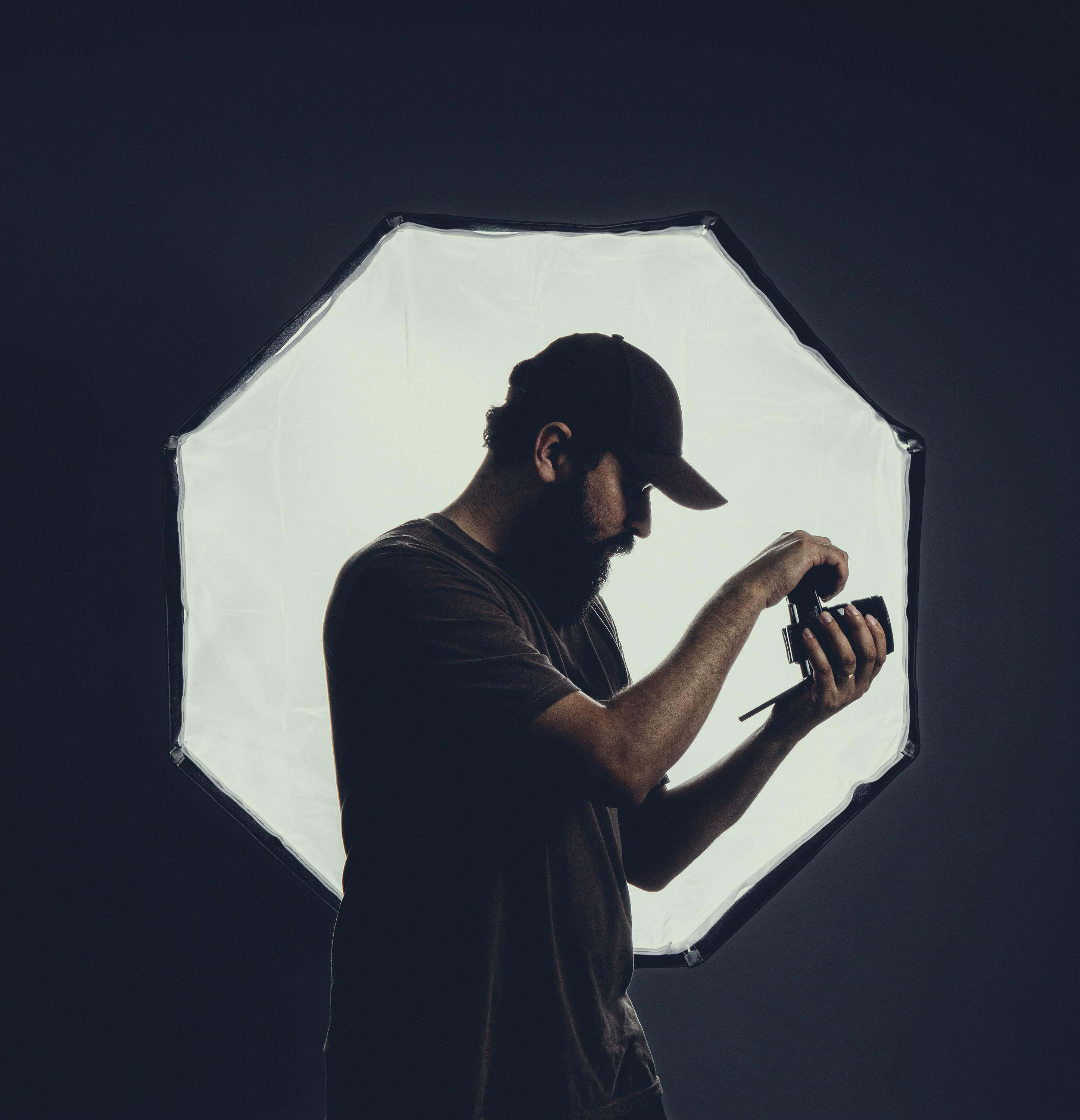 Free Silhouette of a photographer adjusting a camera against a bright softbox in a studio. Stock Photo