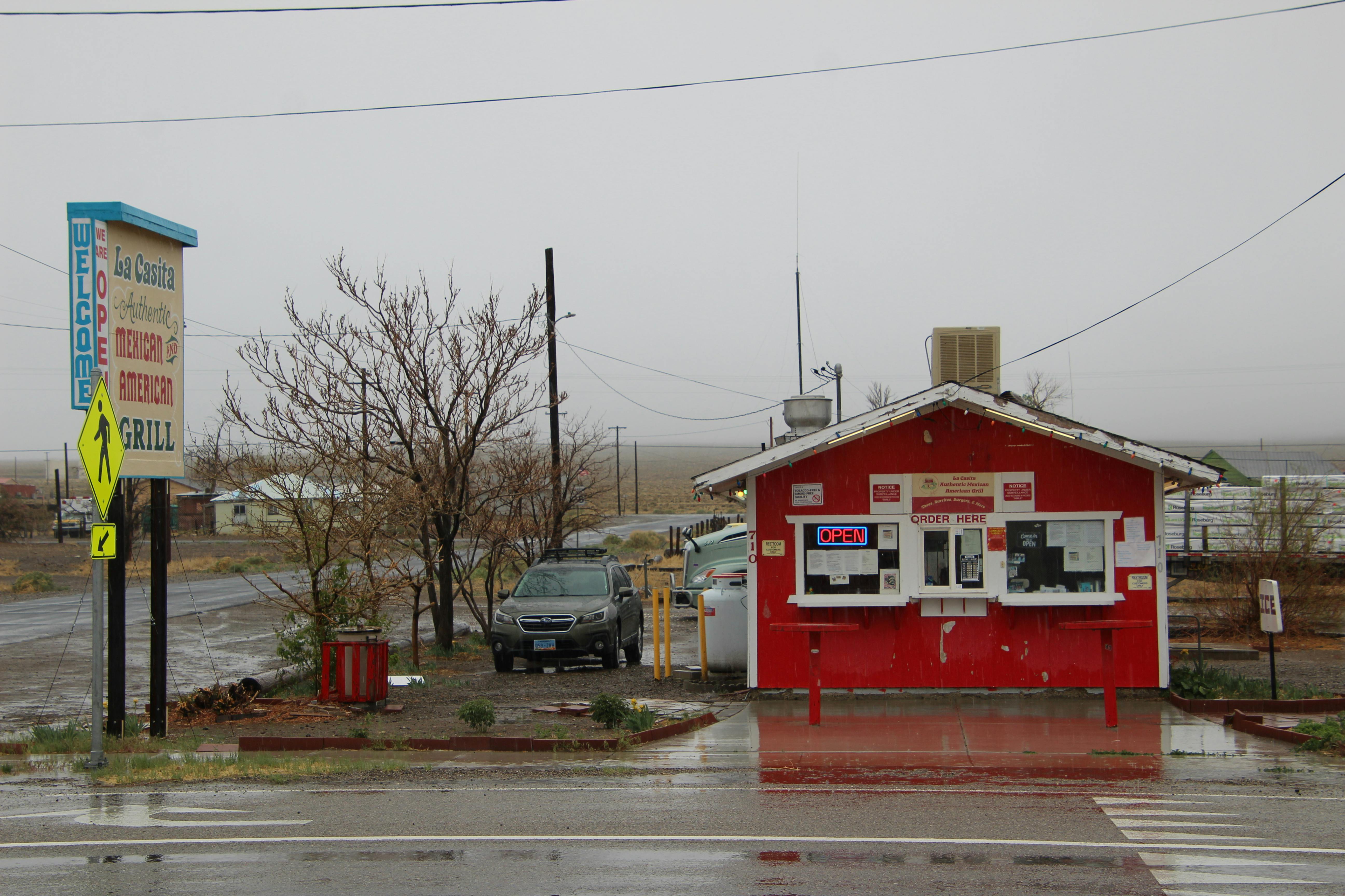 A small red grill shack stands on a rainy day, with an open sign and empty surroundings.