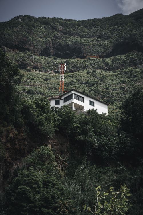 Free White house nestled in a lush green mountain landscape with a telecom tower under cloudy skies. Stock Photo