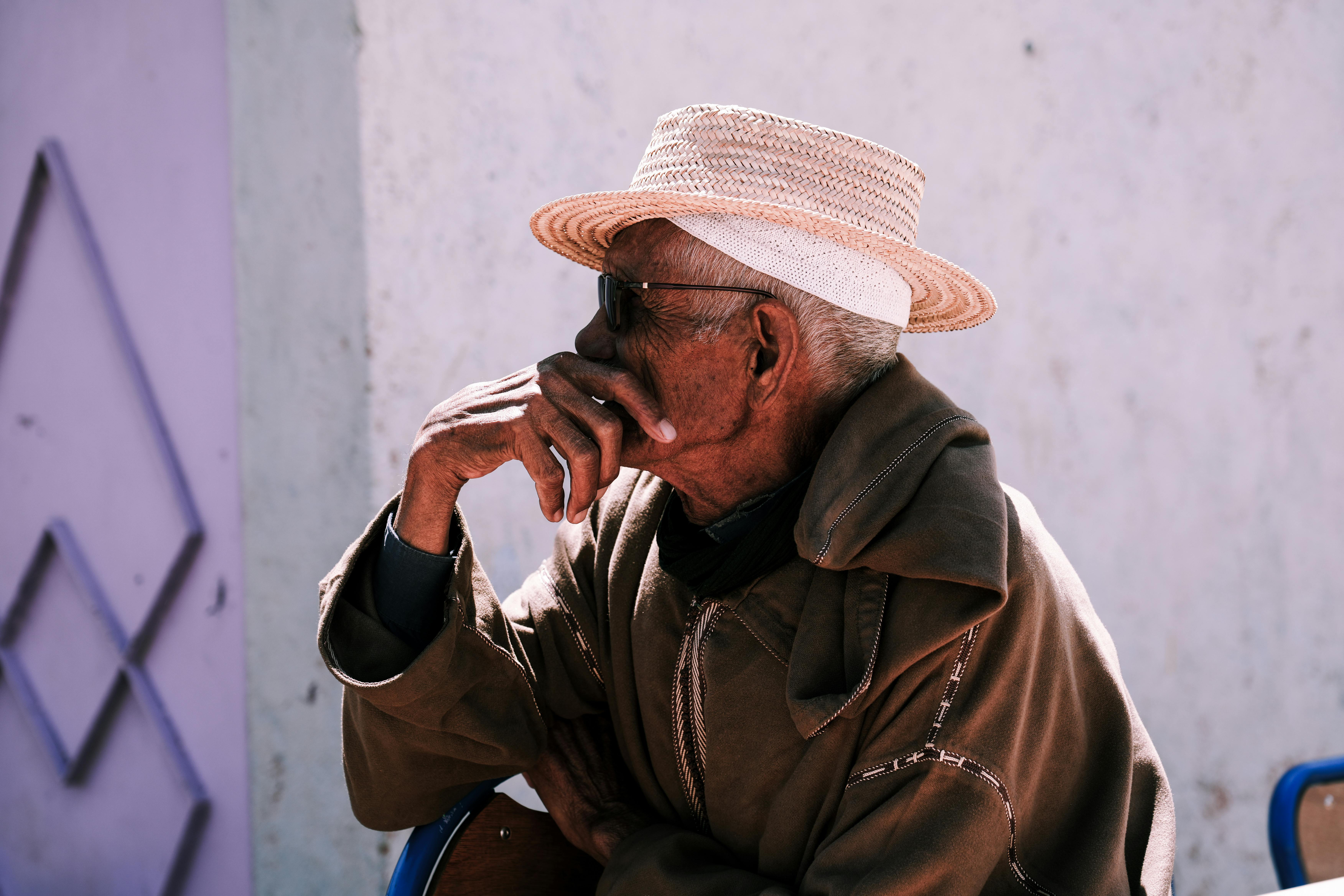 A reflective elderly man wearing a straw hat and coat sits outdoors against a textured wall.