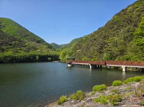 A beautiful spring landscape featuring a serene mountain lake with a wooden walkway surrounded by lush greenery.