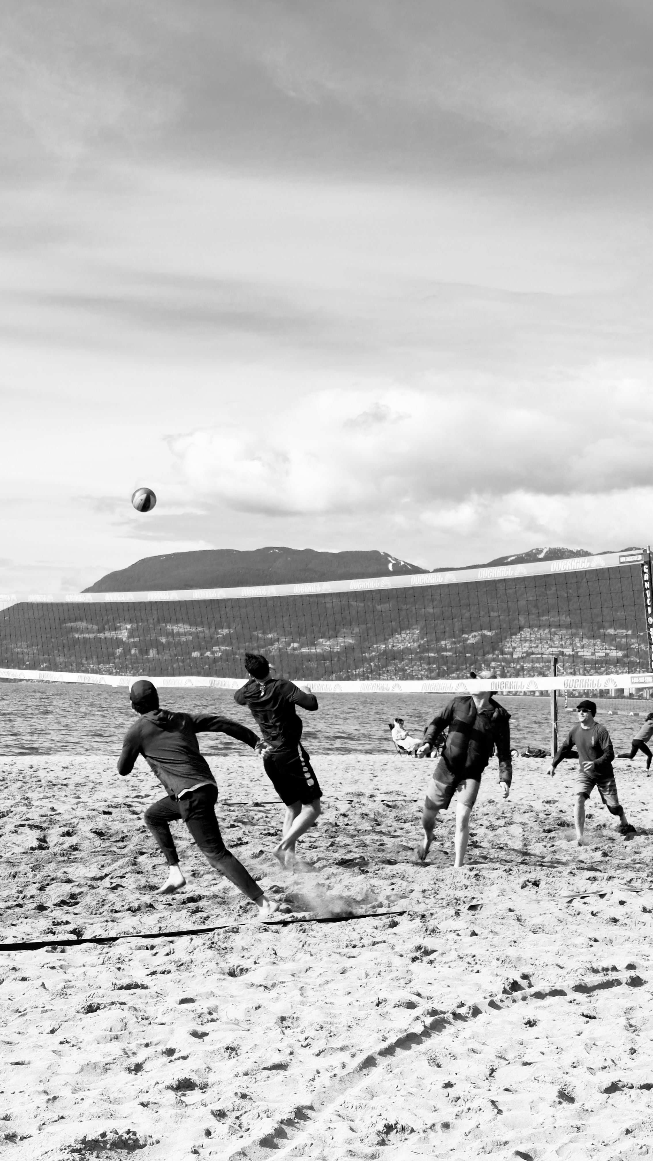 Black and white photo of a beach volleyball game with mountains in the background.