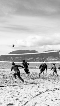 Black and white photo of a beach volleyball game with mountains in the background.