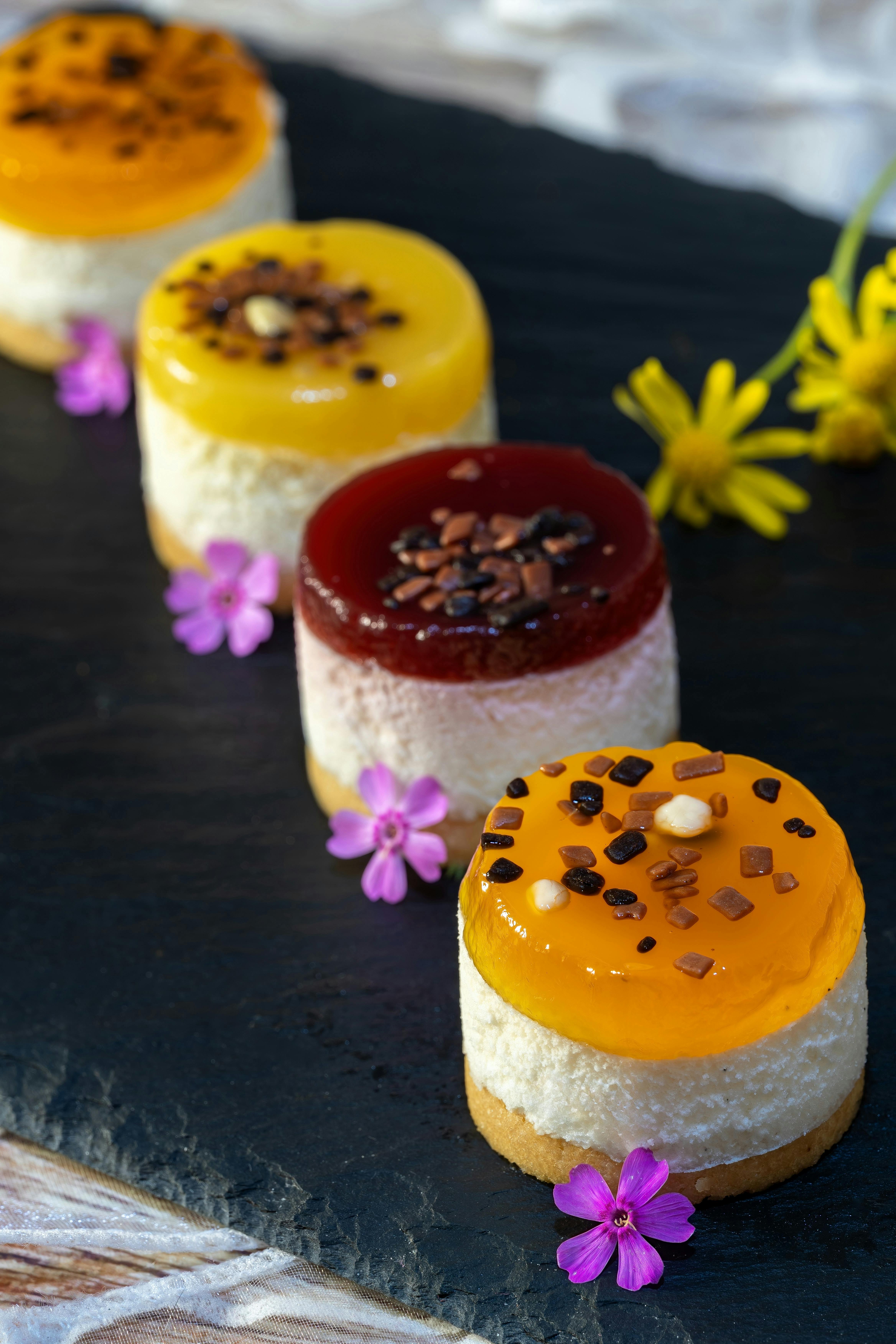 Vibrant mini cheesecakes topped with assorted fruit jelly and flowers on a slate board, ideal for summer desserts.