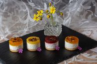 Assorted mini fruit cheesecakes with vibrant toppings presented on a slate board with flowers.
