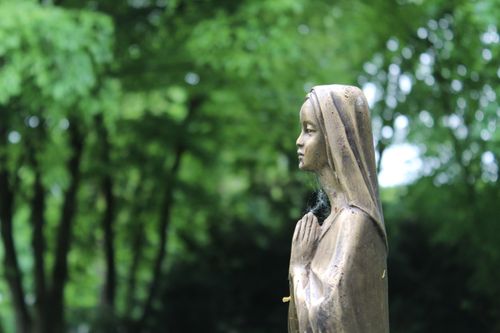 Free A bronze Marian statue in prayer set against a lush green forest background in Augsburg, Germany. Stock Photo
