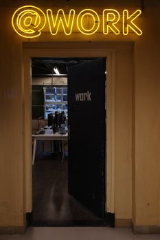 Open office door under a neon '@work' sign leading to a workspace with desks and computers.