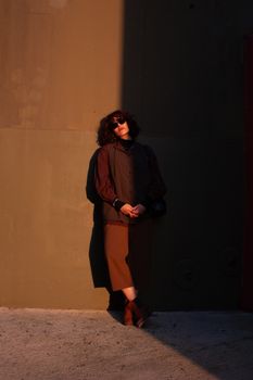 Dramatic sunlight casts shadows on a woman in brown fashion attire against a minimalist wall.
