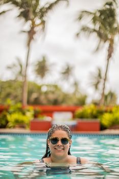 Young woman smiling in a tropical resort pool surrounded by palm trees and sunshine.