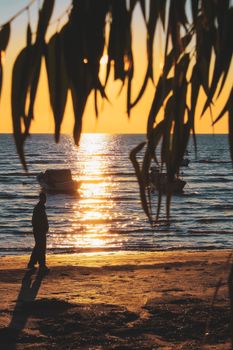Silhouette of a person walking along a beach at sunset with boats in the distance.