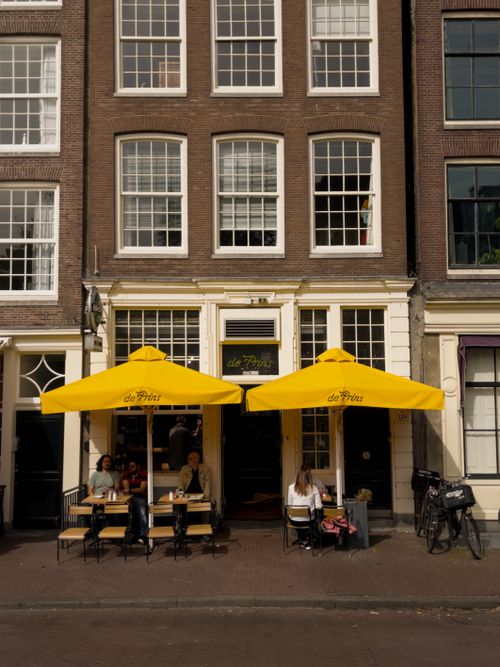 Free Street view of a cozy café in Amsterdam with yellow umbrellas and people enjoying a sunny day. Stock Photo
