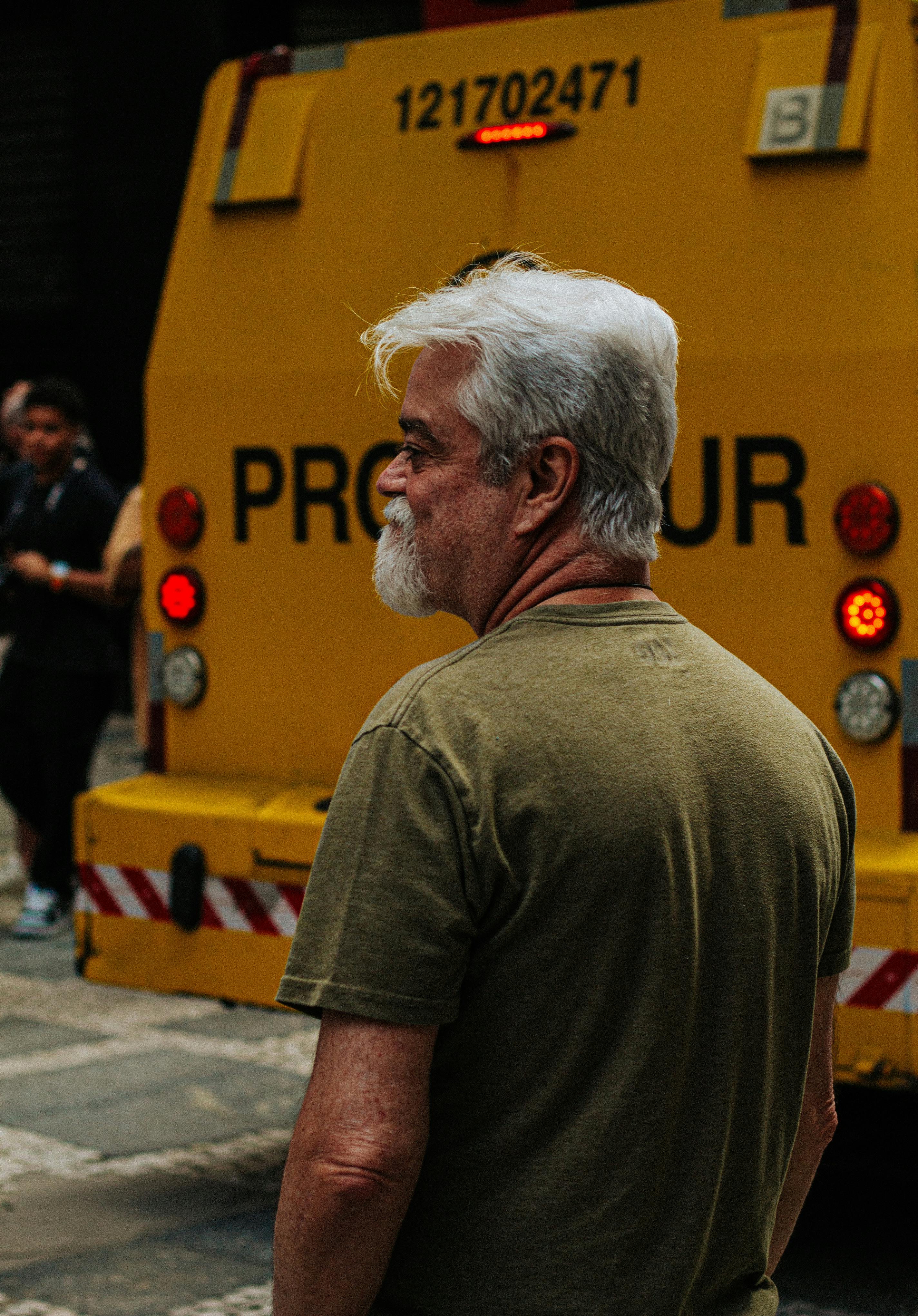 A senior man with white hair stands near a yellow vehicle on an urban street.