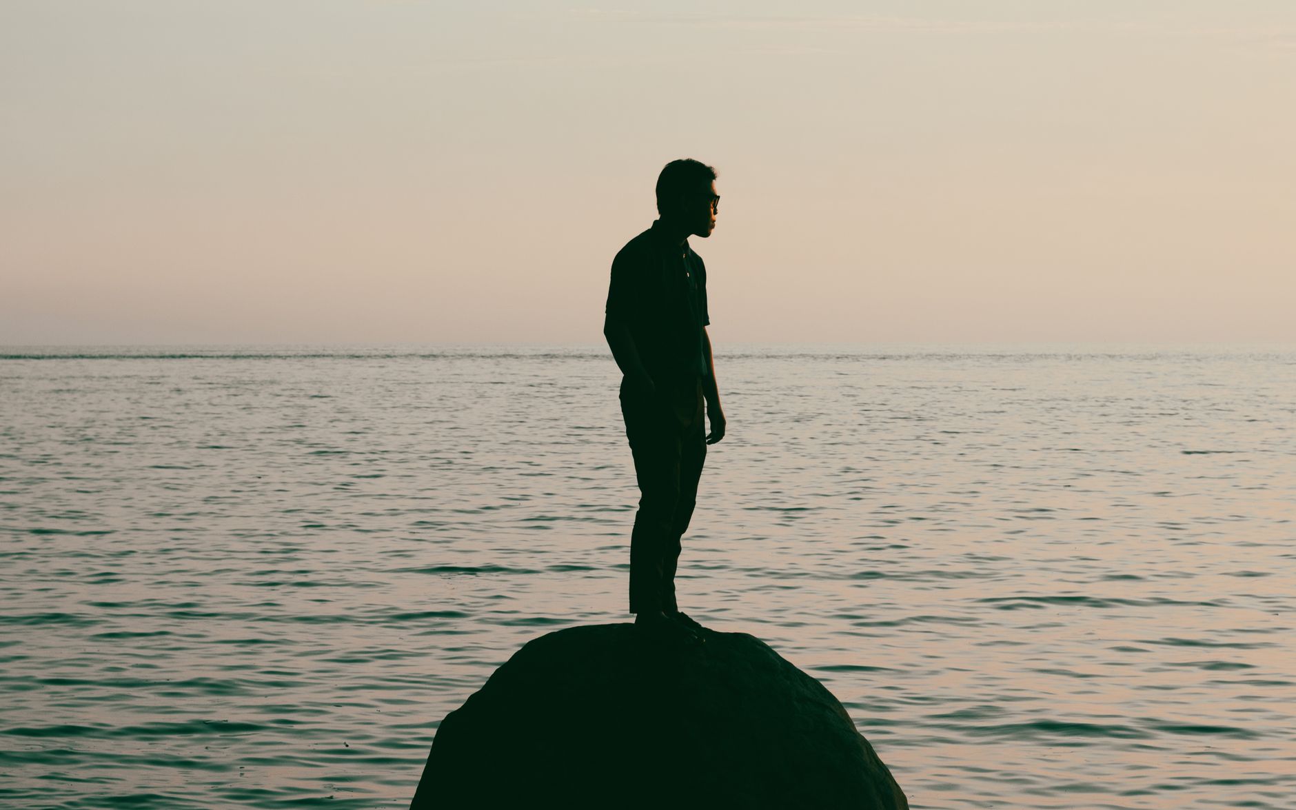 A man stands on a rock by the ocean at sunset, creating a serene silhouette in Gorontalo, Indonesia.