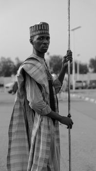 Black and white portrait of a man in Nigerian attire holding a staff outdoors.