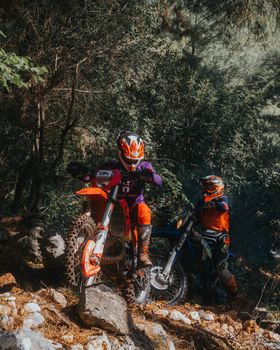 Two motorcyclists navigate rocky terrain in a dense forest during an off-road adventure.