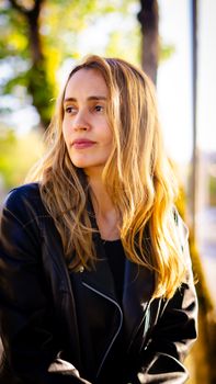 A young woman with blonde hair in a leather jacket enjoys a sunny day outdoors.
