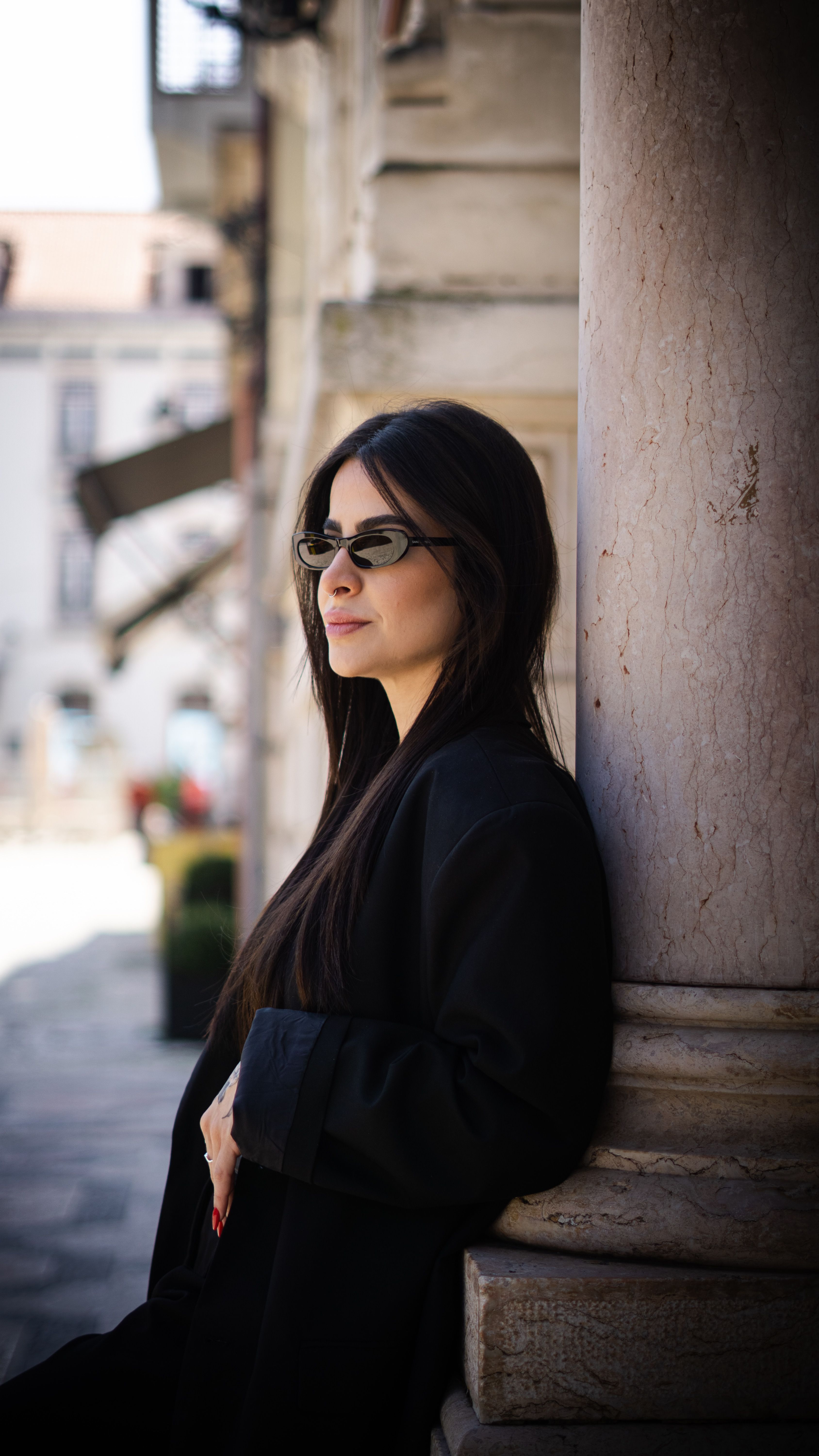 Stylish woman wearing sunglasses, posing against a stone pillar in an urban outdoor setting.