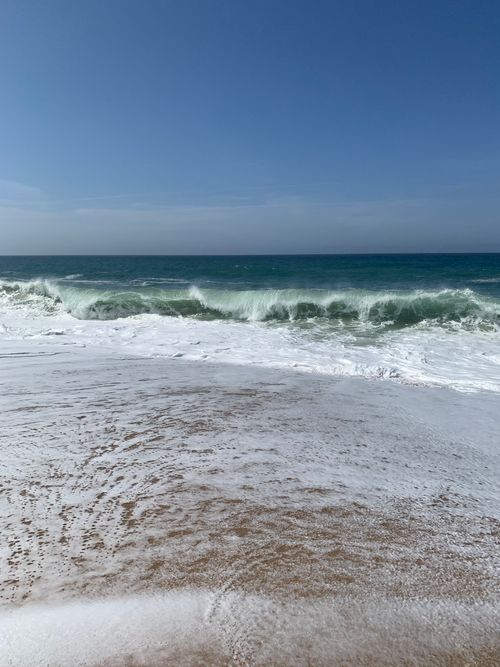 Free Serene beach setting with waves crashing on Nazaré's sandy shore under clear skies. Stock Photo