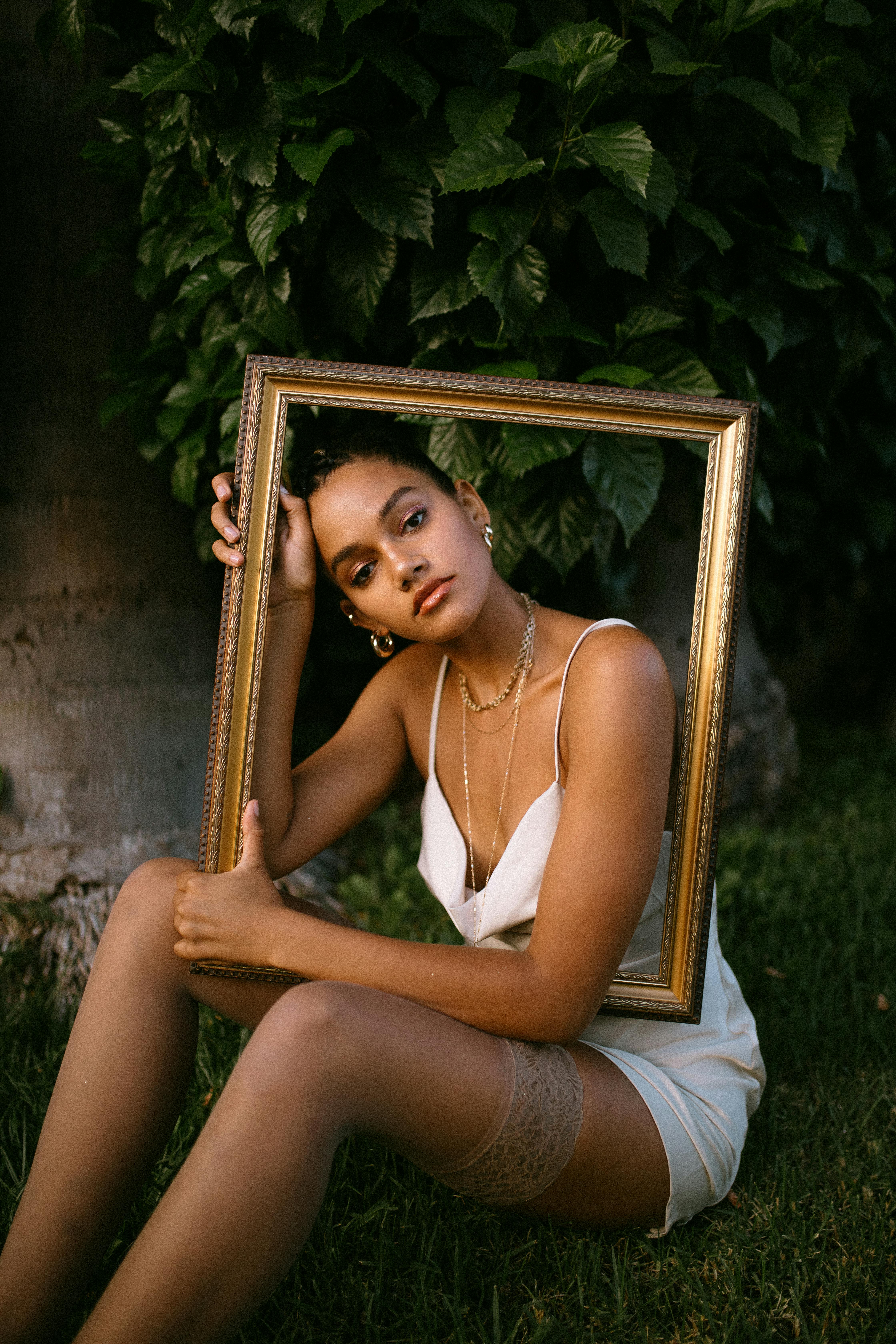 woman in white spaghetti strap top sitting on grass holding brown painting frame