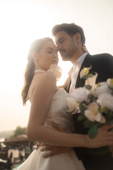 Elegant couple embraces outdoors with bouquet, soft backlight.