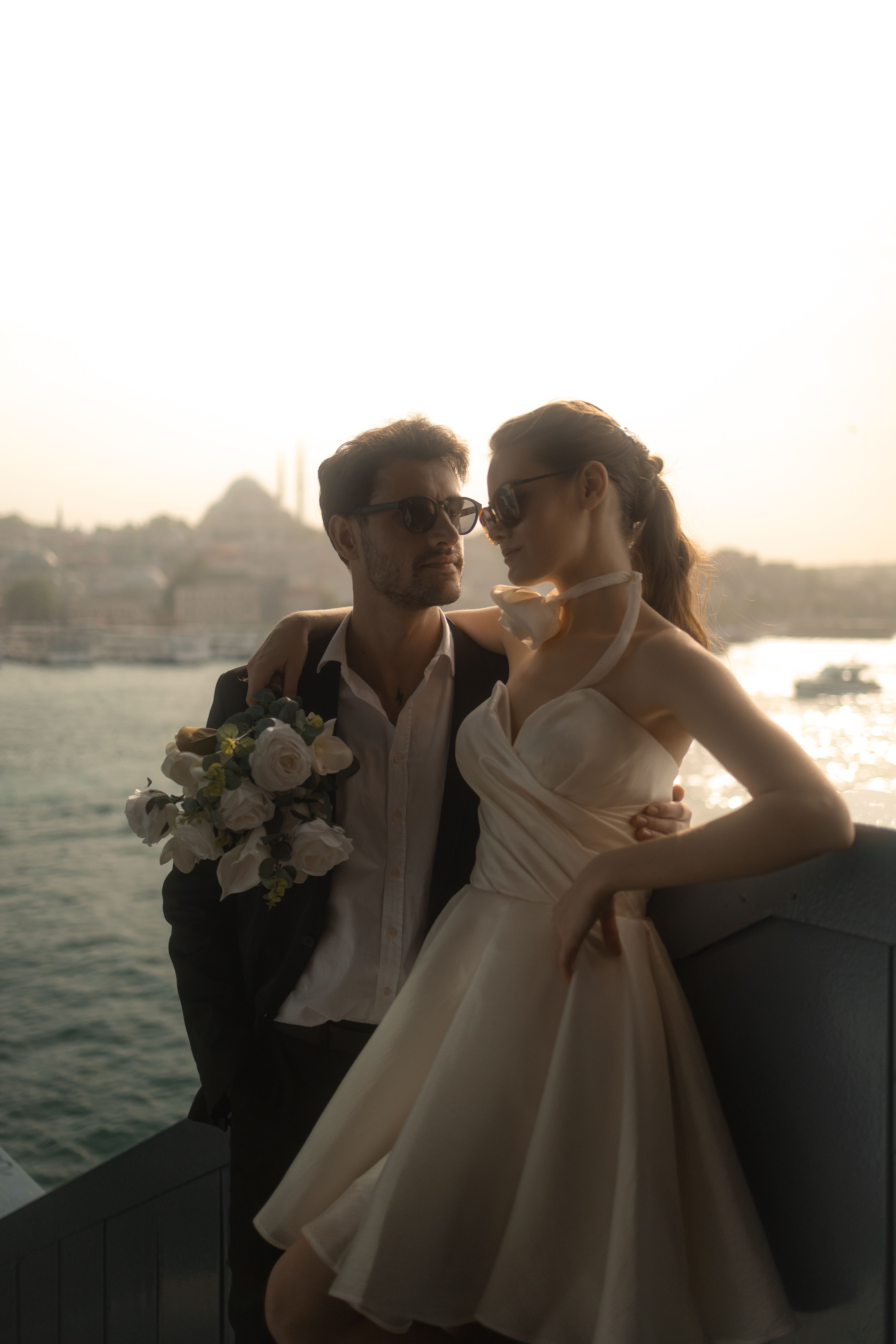 Stylish couple embraces by a scenic waterfront at sunset, exuding romance.