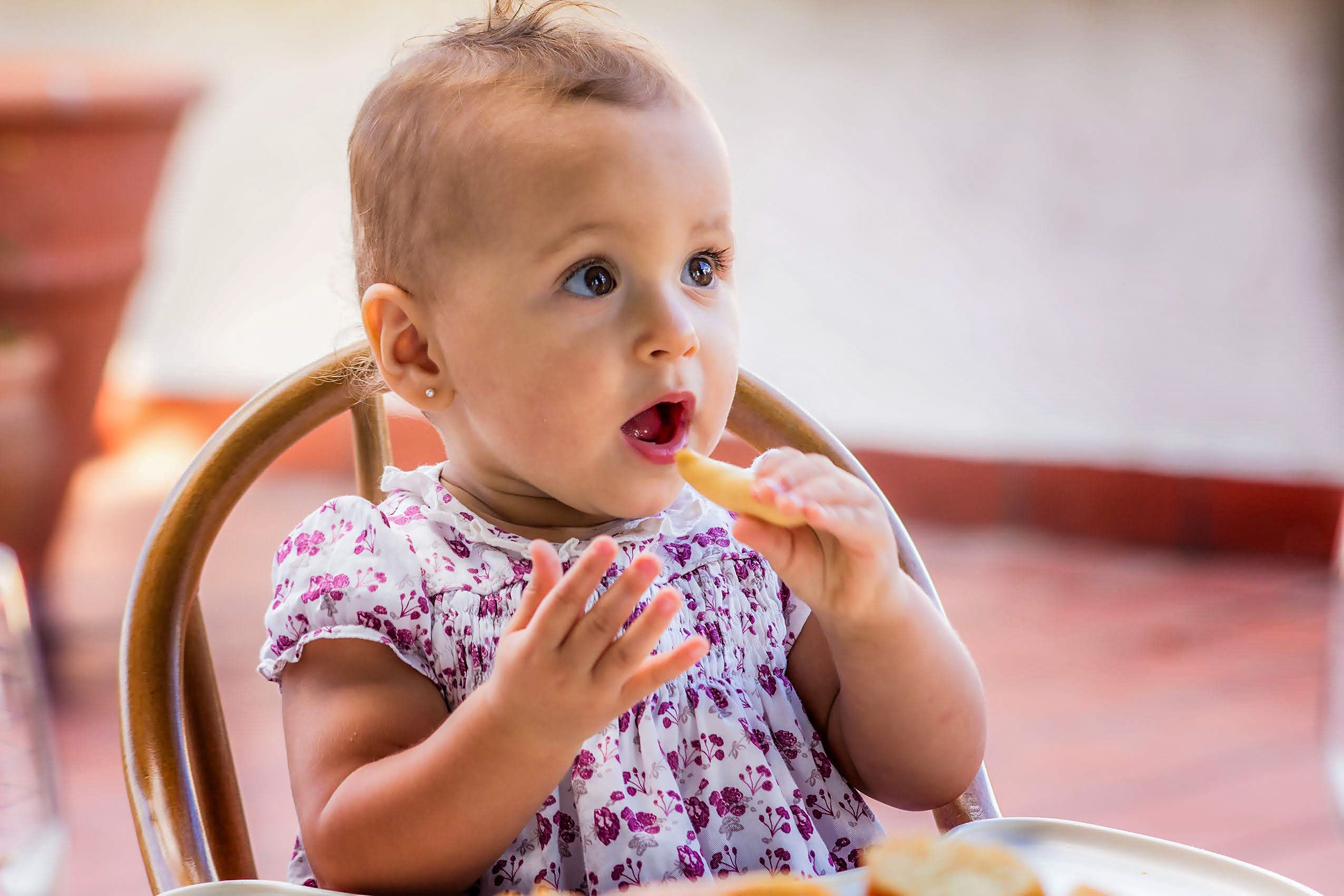 Cute baby girl in floral dress munching on a snack, enjoying a sunny day outside.