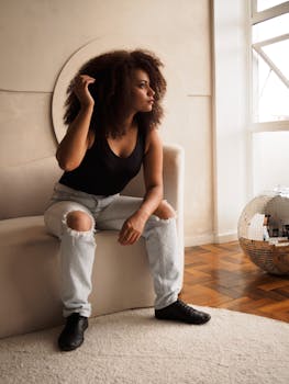 Captured indoors, a woman with curly hair in casual attire sitting thoughtfully on a couch.